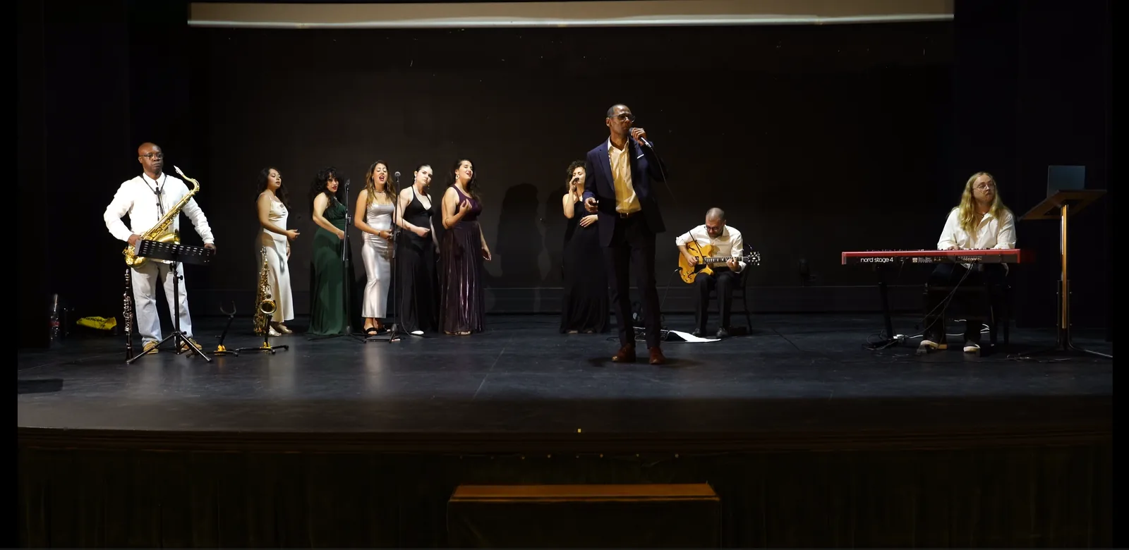 El elenco de Soul & Silence sobre el escenario durante un ensayo: JC Brown cantando, las coristas, saxo, guitarra y piano.